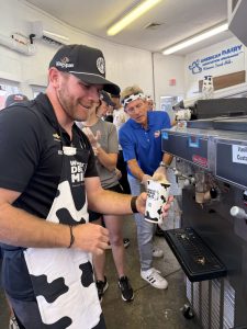 Man in a cow-print apron pours milk from a large espresso-style machine at a dairy booth, smiling at the cup he’s filling.