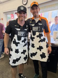 Two men in cow-pattern aprons and orange caps smile for the camera indoors.